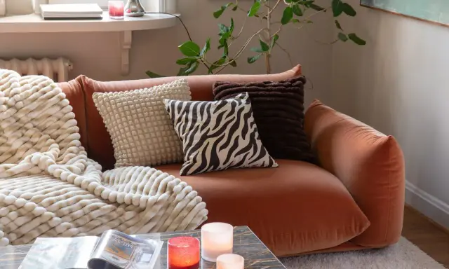 En orange soffa med flera kuddar och en mjuk pläd. Fina dekorationer som ljus och en tidning på soffbordet skapar en mysig atmosfär.