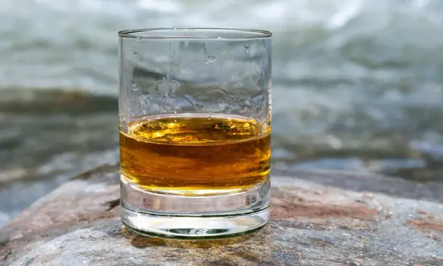 Ein Glas goldener Whisky steht auf einem Stein am Flussufer. Die ideale trinktemperatur für diesen edlen Tropfen.