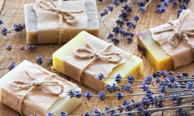 Quattro saponette fatte in casa, avvolte in carta e legate con spago, giacciono su un tavolo di legno rustico, circondate da rametti di lavanda profumata.