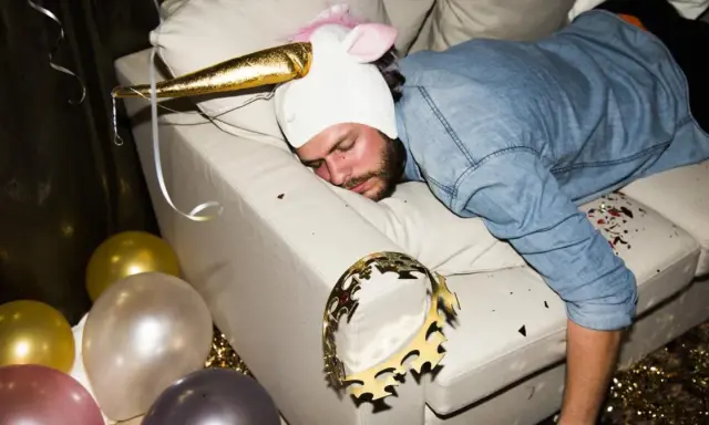 Mężczyzna w stroju jednorożca śpi na kanapie po imprezie. Balony i konfetti sugerują, że to może być sposób na to, jak pozbyć się kaca.