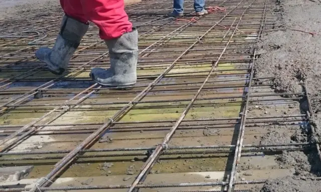 Pracownicy w gumowcach stąpają po siatce zbrojeniowej, przygotowując zbrojenie stropu do zalania betonem.
