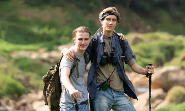 Dwóch młodych mężczyzn, gotowych na trekking, z plecakami i kijkami, w otoczeniu zieleni i skał.