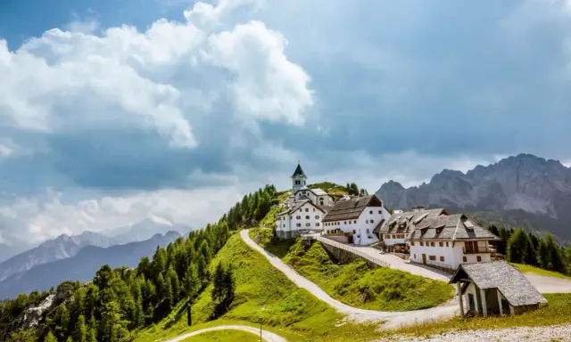 Un borgo incantevole in montagna, perfetto per le tue vacanze in Friuli. Case bianche, una chiesa e sentieri verdi sotto un cielo nuvoloso.