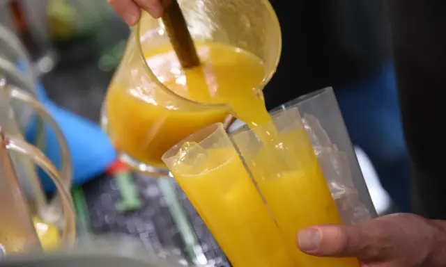 Ein Mann gießt leuchtend gelben Saft, vielleicht Poncha Madeira, aus einer Glaskaraffe in zwei Gläser mit Eiswürfeln.