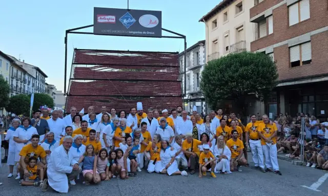 Grupo posa en la fiesta longaniza Graus, con un cartel de "Los Tres Sabores de la Longaniza de Graus" y una estructura de madera con embutidos.