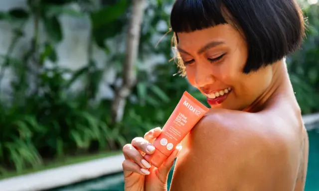 Une femme souriante applique une crème solaire SPF 50+ sur son épaule. Le tube indique "Très haute protection", le SPF 50 signification est une protection optimale.
