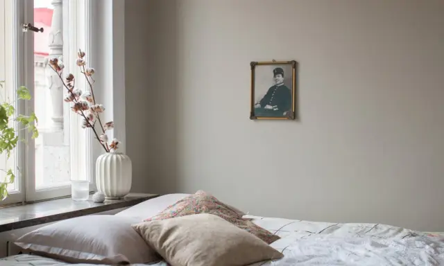 En säng med flera kuddar i olika nyanser av beige och mönster. En gammal porträttbild hänger på väggen.