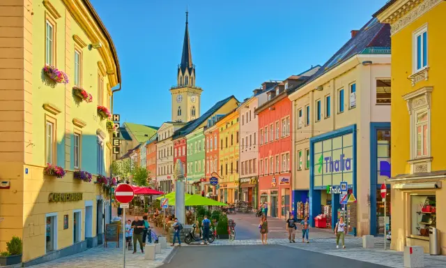 Via colorata di una città austriaca più vicina all'Italia, con edifici vivaci, una torre dell'orologio e gente che passeggia.