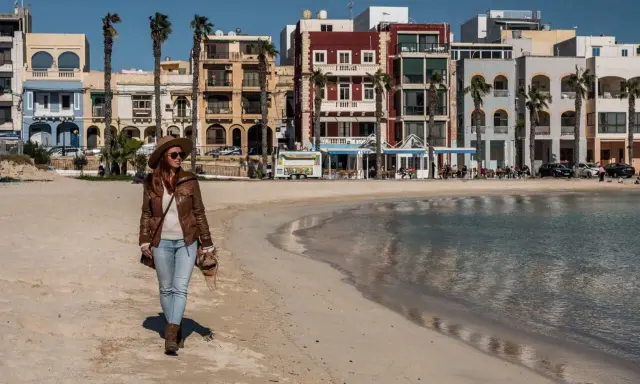 Kobieta spaceruje po piaszczystej plaży w pretty bay Malta, w tle kolorowe budynki i palmy.