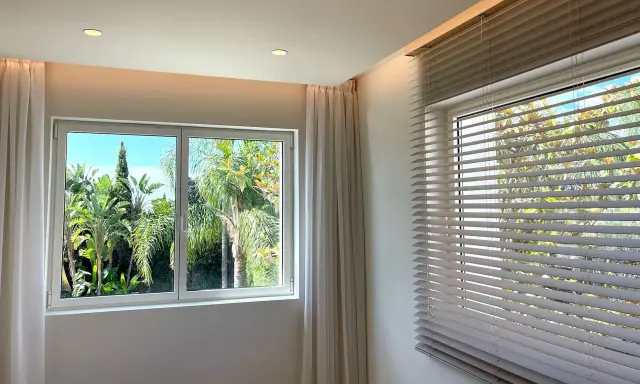 Dos tipos de apertura de ventanas: una corredera con cortinas y otra con persianas venecianas, ambas con vistas a un exuberante jardín tropical.