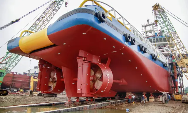 Gros navire rouge et bleu sur des supports, avec des hélices visibles. Des grues et des ouvriers sont présents. On aperçoit des anodes sur la coque, protégeant contre la corrosion, qu'elles soient anode + ou -.