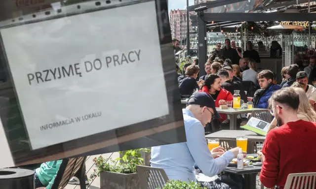 Ogłoszenie "Przyjmę do pracy" na tle ogródka restauracyjnego z ludźmi przy stolikach.