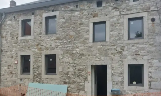 Maison ancienne en cours de rénovation, avec des murs en pierre brute et de nouvelles fenêtres. Le projet de construire en pierre continue.