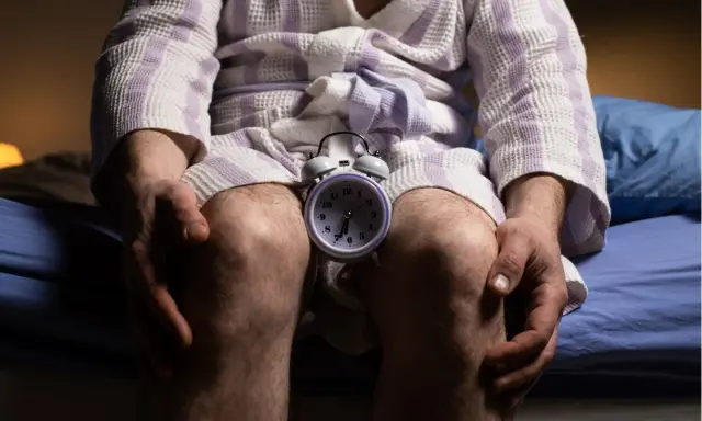Mężczyzna w szlafroku siedzi na łóżku z budzikiem między kolanami. Czas na rozmowę o andropauzie a seksualności.