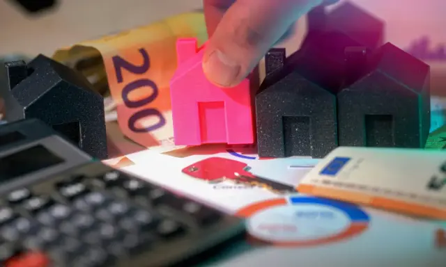 Petites maisons, billets de banque et calculatrice symbolisant le déblocage des fonds pour un projet immobilier. Une main place une maison rose.