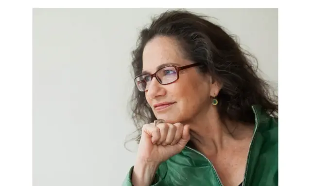 Susan Neiman, met een denkbeeldige blik, draagt een groene jas en een bril.