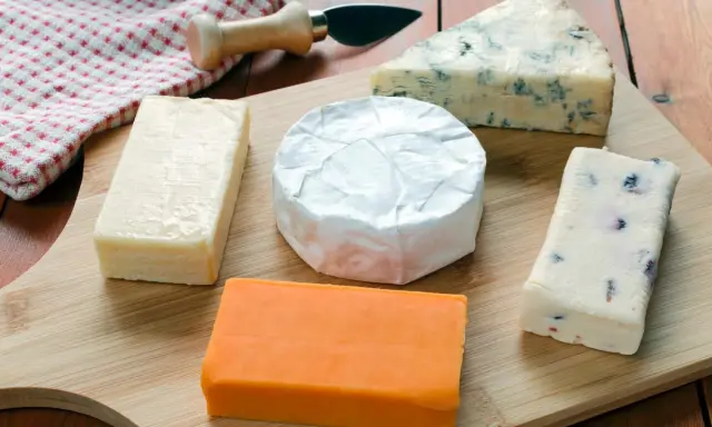 Verschiedene Käsesorten auf einem Holzbrett. Warum ist Cheddar orange? Die natürliche Farbe kommt von Beta-Carotin, das Kühe über Gras aufnehmen.