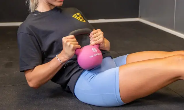 Frau macht ein **Kugelhantel Training** auf dem Boden, hält eine pinke Kettlebell und trainiert ihre Bauchmuskeln.
