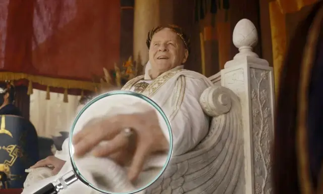 Ein Mann mit Lorbeerkranz, der an alle Kaiser Roms erinnert, sitzt auf einem Thron. Eine Lupe enthüllt Details seiner Hand.