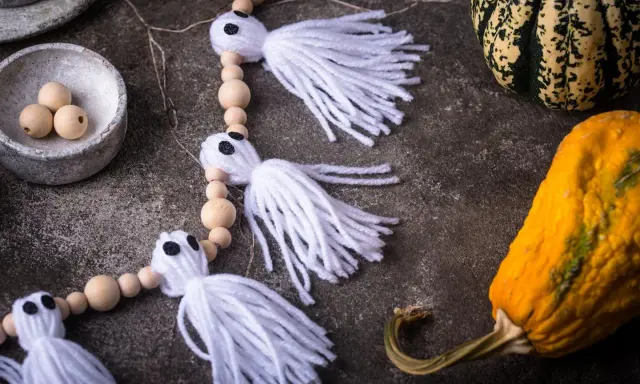 Niedliche Geister-Girlande zum Halloween basteln, mit Holzperlen und weißen Fransen. Daneben liegen bunte Kürbisse.