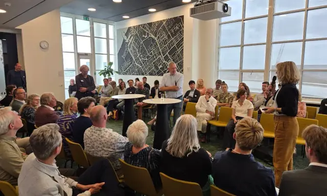 Mannen en vrouwen luisteren aandachtig naar een spreker, mogelijk over ruimtelijke ordening, in een zaal met een grote stadskaart aan de muur.