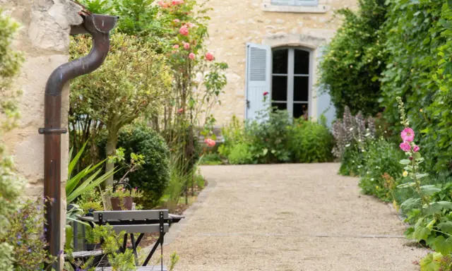 Un chemin de gravier serpente à travers un jardin anglais romantique, bordé de roses roses et de verdure luxuriante. Une maison en pierre avec des volets bleus se profile à l'arrière.