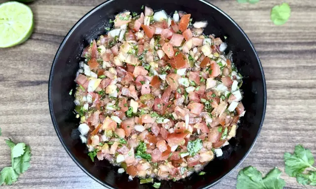 Świeże, kolorowe pico de gallo w czarnej misce, z limonkami i kolendrą na drewnianym tle.