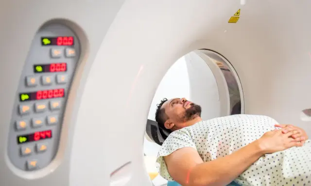 Pacjent leży w aparacie do tomografii komputerowej, przygotowując się do badania. Obok widoczny panel sterowania z cyfrowymi wskaźnikami.
