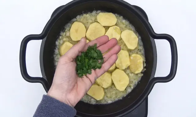 Manos añaden perejil picado a patatas en rodajas sobre cebolla sofrita en una sartén. ¡El inicio de unas deliciosas patatas en salsa verde!