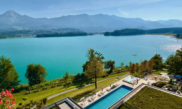 Turkusowe wody Ossiacher See lśnią w słońcu, a w oddali widać góry. Obok basenu i jacuzzi rozciąga się zielony trawnik z leżakami.