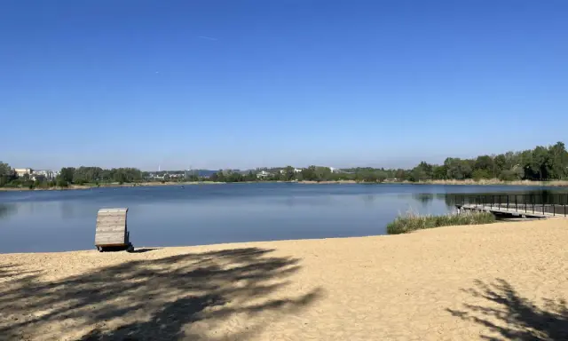 Plaża bagry z drewnianą przebieralnią i pomostem, w tle jezioro i zieleń.