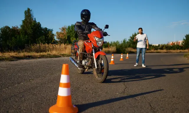 Persona con casco y equipo de protección conduce una moto naranja en un circuito de prácticas. Un instructor observa.