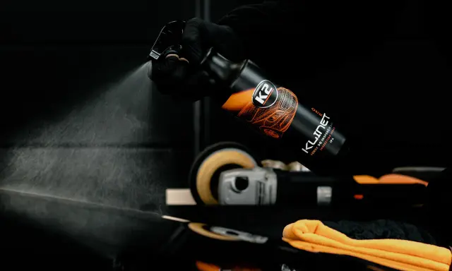 Ręka w czarnej rękawiczce rozpyla płyn K2 Klinet, środek do inspekcji lakieru z alkoholem IPA. W tle polerka i pomarańczowa mikrofibra.