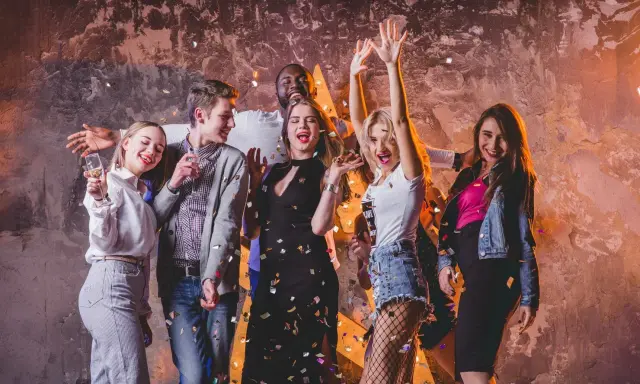 Grupa młodych ludzi świętuje, sypiąc konfetti. Ich stroje to świetna inspiracja, jak się ubrać na dyskotekę: sukienki, jeansy i błyszczące dodatki.