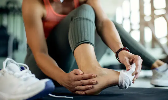 Kobieta w sportowym stroju masuje bolącą kostkę, obok leżą buty sportowe.