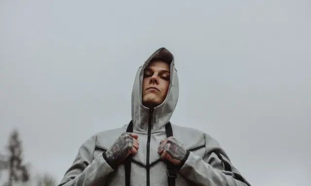 Młody mężczyzna w szarej bluzie z kapturem, z tatuażami na dłoniach, wygląda na zamyślonego. Może to symbol walki z chorobą, jak szpaku choroba, która go trapi.
