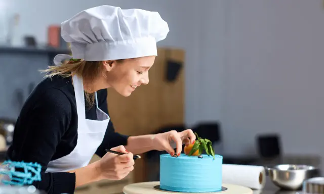 Uśmiechnięta cukierniczka w białym czepku i fartuchu dekoruje błękitny tort. Jej cv to pasja i precyzja w tworzeniu słodkich arcydzieł.