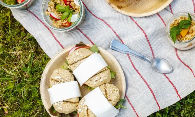 Idée pique-nique : petits pains garnis, salades en bocaux et muffins sur une nappe rayée, posée sur l'herbe.