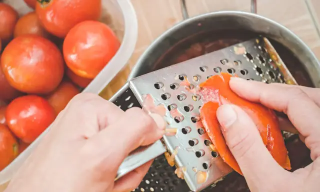 Ręce trą pomidora na tarce, przygotowując go do sosu. Obok stoi miska pełna świeżych pomidorów.