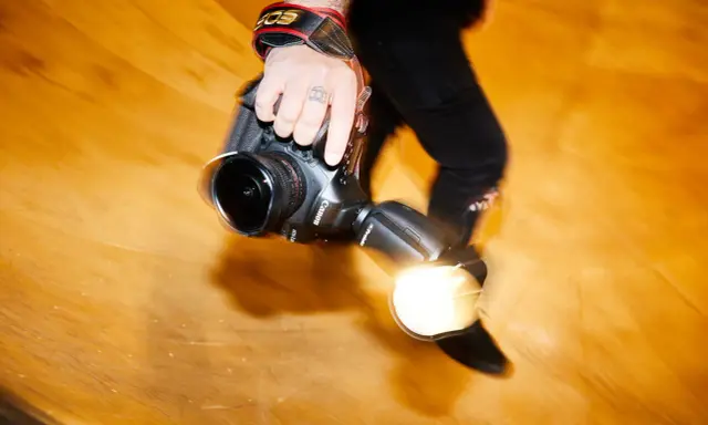 Dłoń fotografa trzyma aparat z lampą błyskową. Długi czas naświetlania tworzy efekt ruchu.