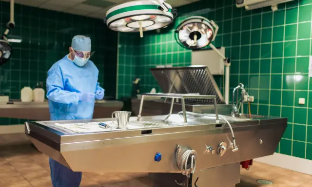 Specjalista w niebieskim fartuchu i masce przygotowuje się do zabiegu tanatopraksji w sterylnym pomieszczeniu z zielonymi kafelkami.