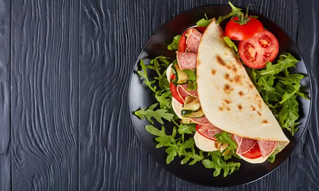 Piadina ripiena con salame, mozzarella, zucchine grigliate e pomodoro, servita su un piatto nero con rucola. Un assaggio della città di Rimini in un ristorante.