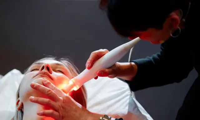 Une esthéticienne utilise un appareil Bloomea pour un soin du visage à la lumière rouge sur une cliente allongée.