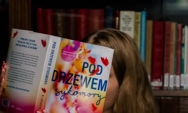 Dziewczyna czyta książkę "Pod drzewem sykomory", ukrywając twarz za okładką. W tle widać regały pełne książek.