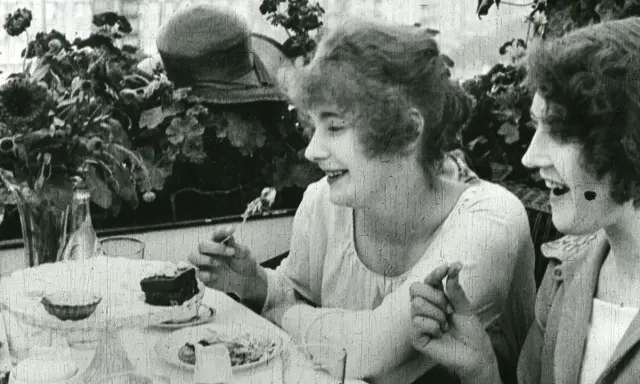 Två kvinnor, en av dem Greta Gustafsson, skrattar vid ett bord med tårta och blommor.