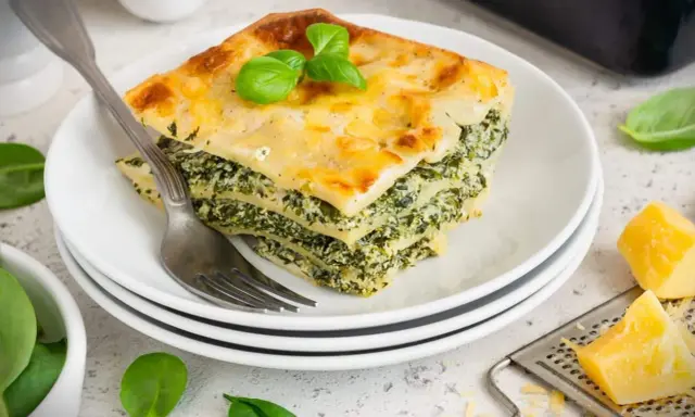 Ein Stück Spinatlasagne mit Käse überbacken, garniert mit Basilikum, auf einem Teller mit einer Gabel.