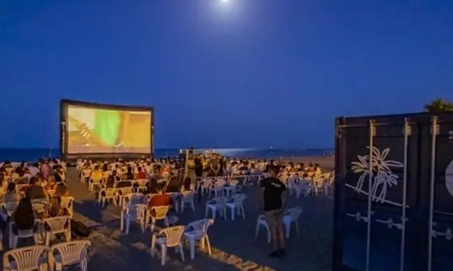 Noche de cine en el parque del cine Málaga. Público sentado en sillas en la playa, viendo una película en una pantalla gigante bajo la luna.