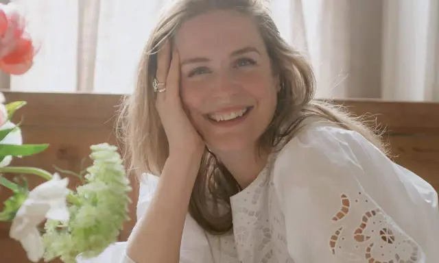 Glad kvinna med blont hår ler mot kameran, med handen mot kinden. En bukett blommor syns till vänster. Kanske ett foto från sofia wood blogg.