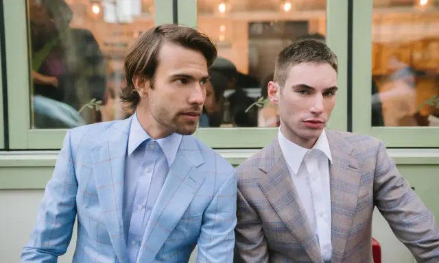 Two men in plaid suits with wide notch lapels sit side-by-side. The man on the left wears a light blue suit, the man on the right a tan suit.