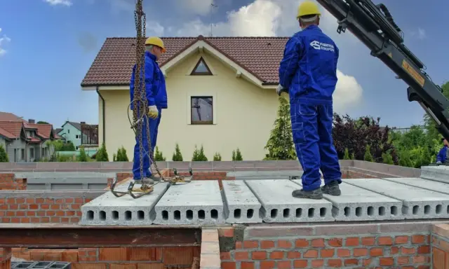 Dwaj pracownicy w niebieskich kombinezonach montują prefabrykowany strop. To kluczowy element budowy domu.